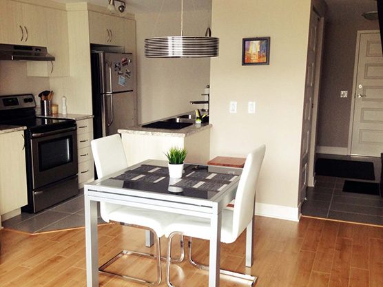 La cuisine d’un appartement : armoires foncées, cuisinière en acier, table carrée avec deux chaises blanches