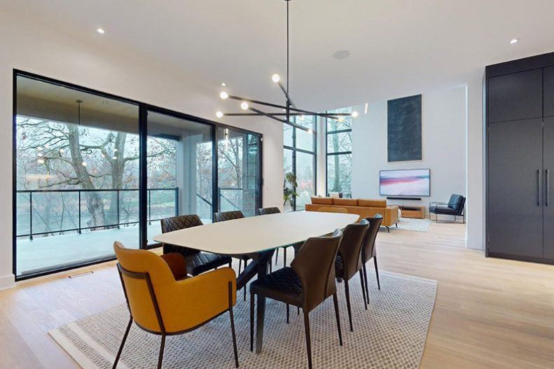 La salle à manger de la maison modèle : table ovale, chaises de cuir cognac, fenêtres sur les arbres