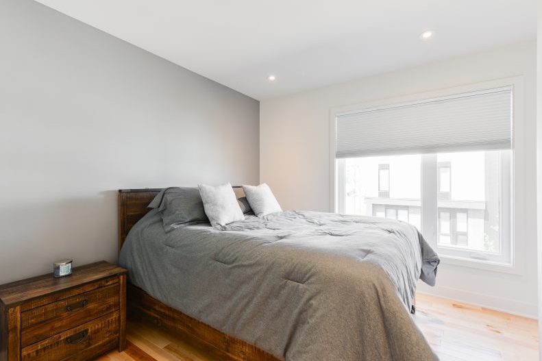 Une chambre aux murs blancs, lit de bois, couette grise, fenêtre sur le voisinage