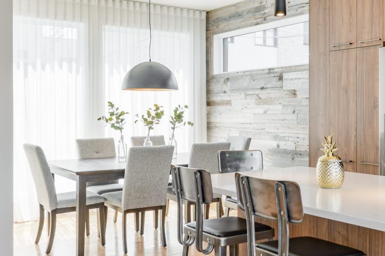 La salle à manger d’une unité : table blanche, chaises grises, mur de planches blanchies, suspension noire